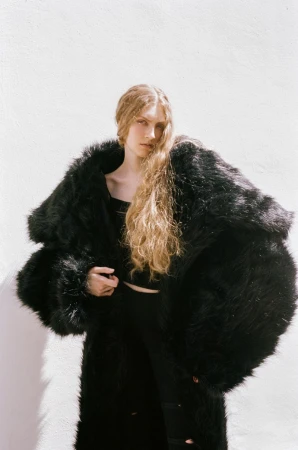 Model with long blonde hair wearing a voluminous black fur coat against a bright white wall