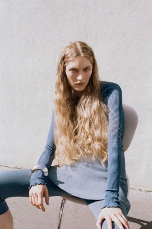 Model with long blonde wavy hair wearing a blue ribbed top and silver cuff seated on a white chair against a textured wall
