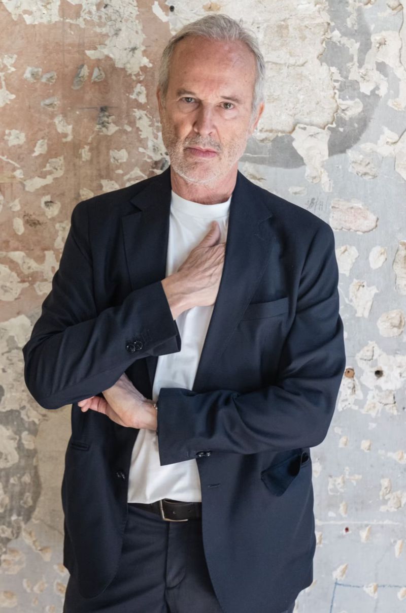 Portrait of artist Erwin Wurm wearing a dark blazer over a white t-shirt against a weathered textured wall background
