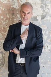 Portrait of artist Erwin Wurm wearing a dark blazer over a white t-shirt against a weathered textured wall background