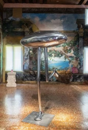 Polished metallic sculpture of a mushroom-shaped object on long thin legs standing in an ornate room with frescoed walls