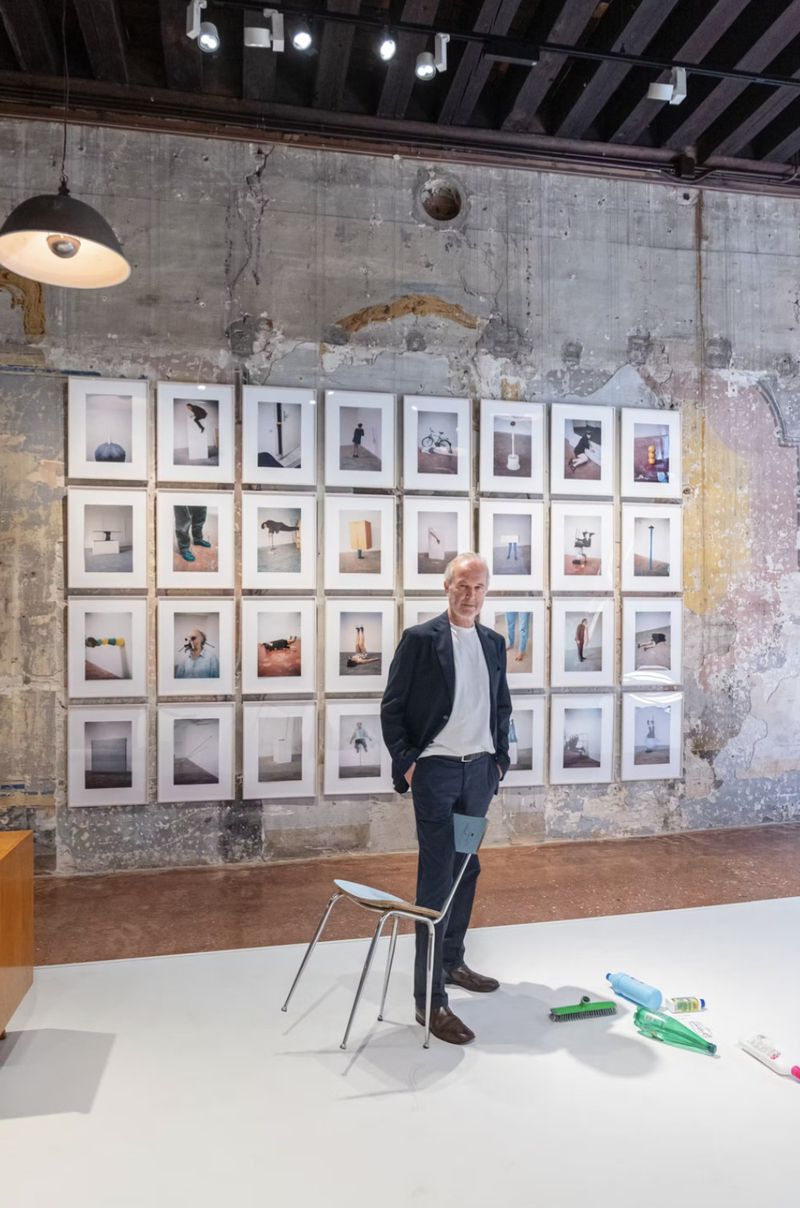 An older man standing in an art gallery before a grid of framed photographs, with a single chair and plastic bottles on the floor.