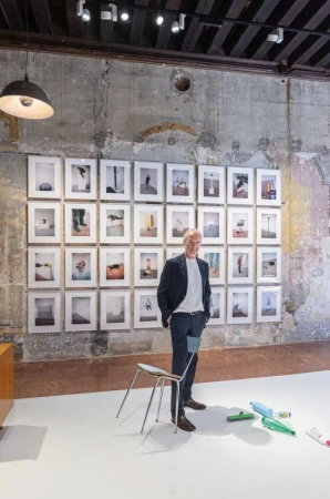 An older man standing in an art gallery before a grid of framed photographs, with a single chair and plastic bottles on the floor.