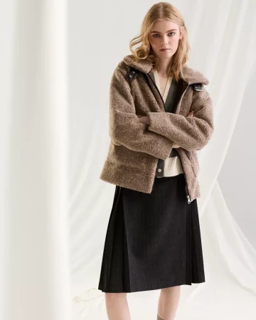 Model with blonde hair wearing a brown textured jacket and black pleated skirt against a white studio backdrop