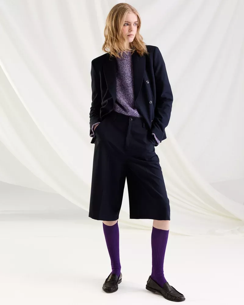 Model wearing a dark blazer, shorts, purple socks, and loafers against a draped white fabric backdrop