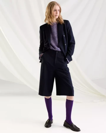 Model wearing a dark blazer, shorts, purple socks, and loafers against a draped white fabric backdrop