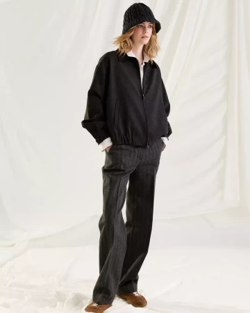 Model wearing a black zip jacket, dark trousers, and a ribbed knit hat against a white draped fabric background