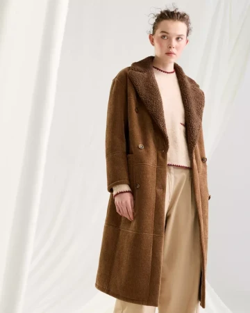 Model wearing a long brown shearling-collar coat and neutral trousers against a white backdrop for Seventy Venezia.