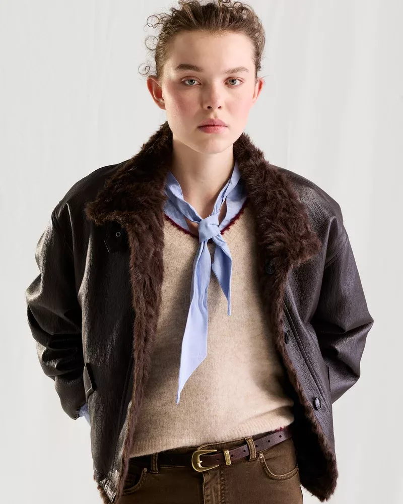 Model in brown shearling jacket over beige sweater and blue scarf against a plain studio background