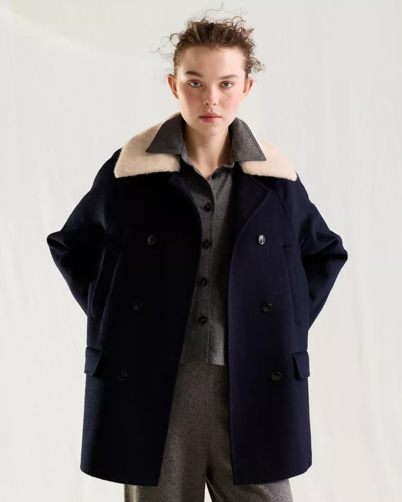 Model wearing a dark double-breasted coat with a light fur collar over grey trousers against a white seamless background