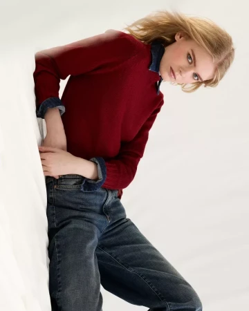 Model with blonde hair wearing a burgundy crewneck sweater over a denim shirt and dark jeans against a white background