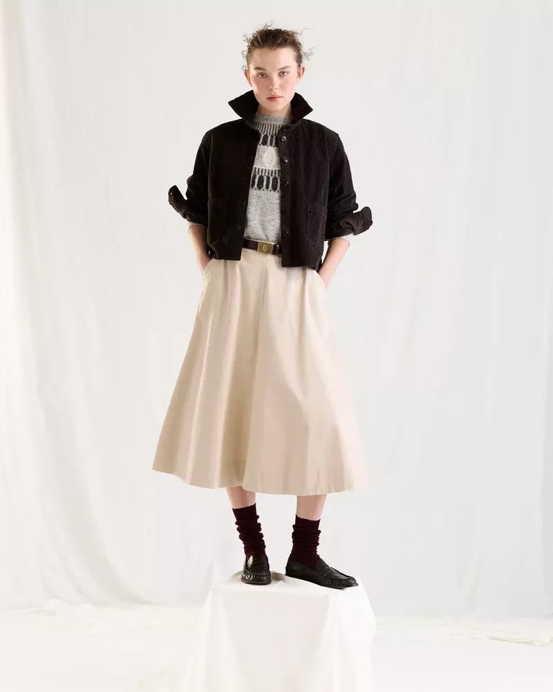 Model standing on a white pedestal wearing a black jacket, patterned knit sweater, cream midi skirt, and dark socks with loafers.