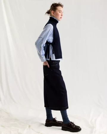 Model in profile wearing a dark navy sleeveless sweater over a light blue button-down shirt and dark skirt against white seamless