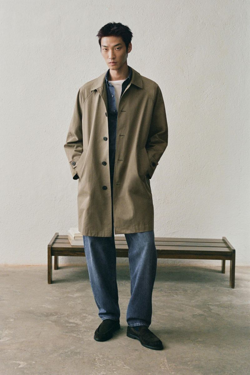 Man wearing a brown trench coat over denim shirt and jeans standing against a textured plaster wall with a wooden bench behind him.