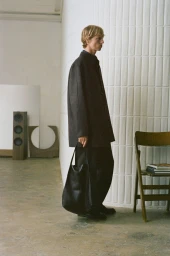 Model in dark jacket carrying a large black leather tote bag standing against a textured white wall in an interior space