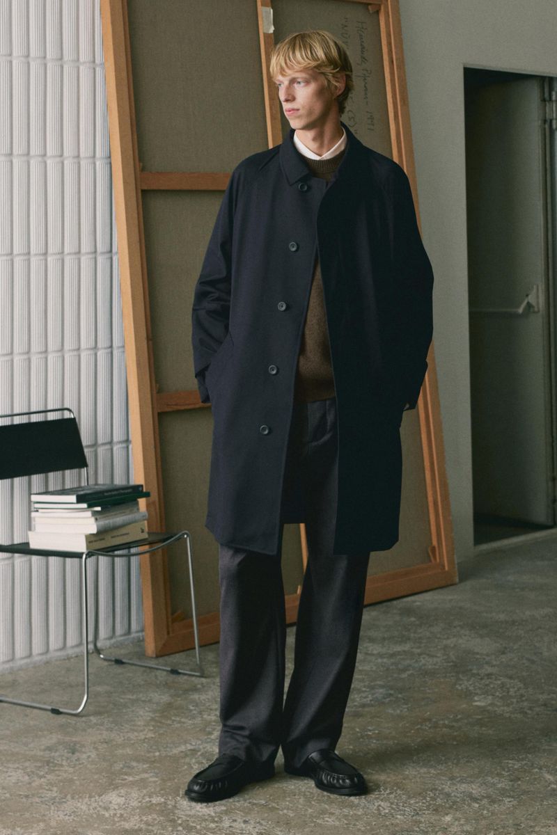 Model standing in a long dark coat and trousers inside an artist studio with large wooden canvases and books.