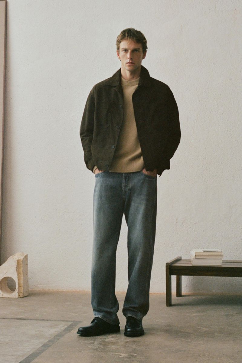 A man wearing a dark jacket, beige sweater, and grey jeans standing in a minimalist room with a low wooden bench.