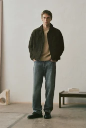 A man wearing a dark jacket, beige sweater, and grey jeans standing in a minimalist room with a low wooden bench.