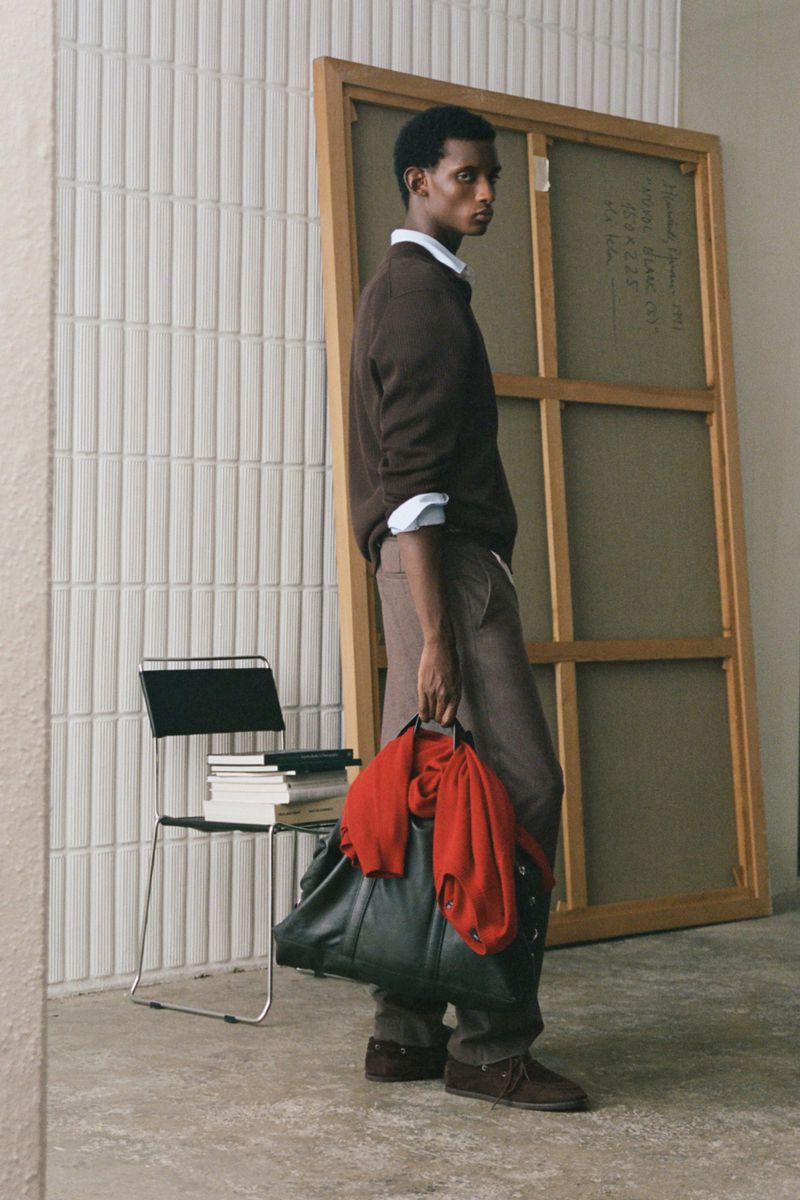 Model standing in brown sweater and trousers holding a large dark bag with a red sweater draped over it