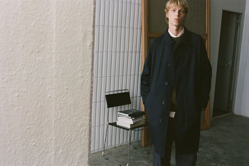 A young male model standing indoors wearing a dark coat and collared shirt next to a chair stacked with books.