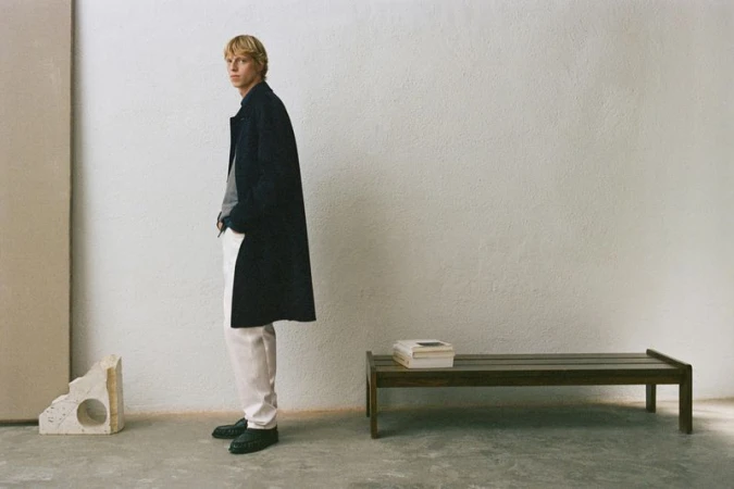 Model wearing dark coat and white trousers standing against a textured wall with a wooden bench