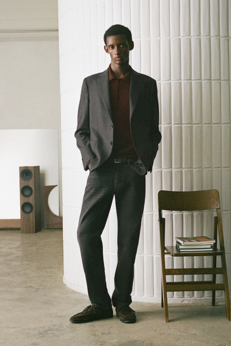 Man wearing a grey suit jacket and trousers posing against a fluted white pillar next to a wooden folding chair