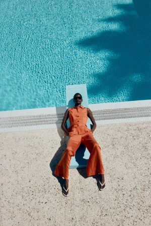 Model in orange sleeveless top and matching trousers lying on a blue diving board by a swimming pool for Zara.