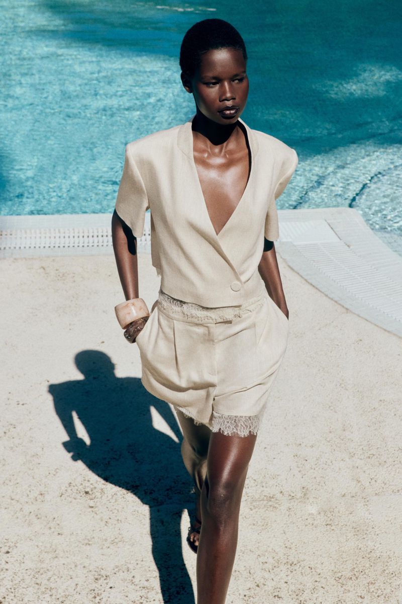 Model Mary Ukech walking outdoors near an outdoor pool wearing a cream cropped blazer and shorts for the Zara project.
