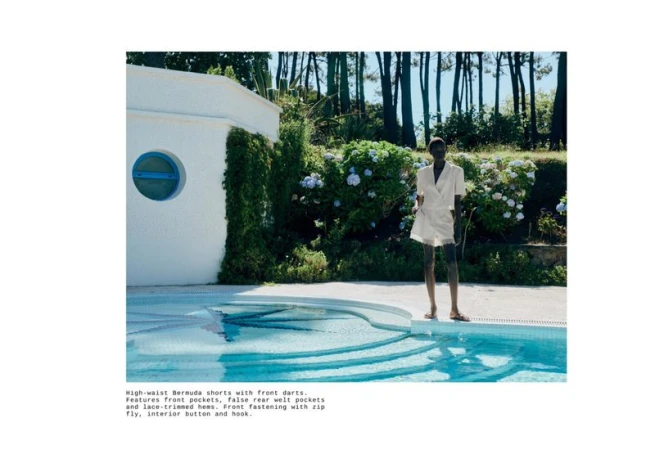 Model standing by an outdoor swimming pool wearing white shorts and a matching top in a sunny garden setting.