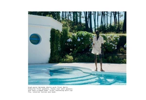 Model standing by an outdoor swimming pool wearing white shorts and a matching top in a sunny garden setting.