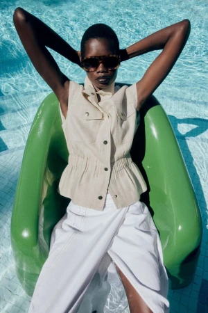 Model wearing tortoiseshell sunglasses and a cream vest seated in a green chair by a pool for Mary X Zara.