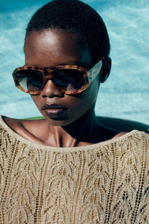 Close-up portrait of a model wearing tortoiseshell sunglasses and a textured metallic gold knit top by the water.