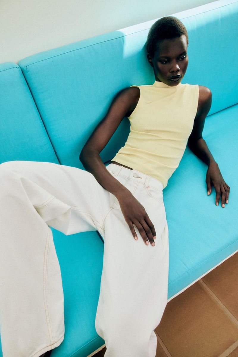 Model wearing yellow sleeveless top and white trousers reclining on bright blue couch