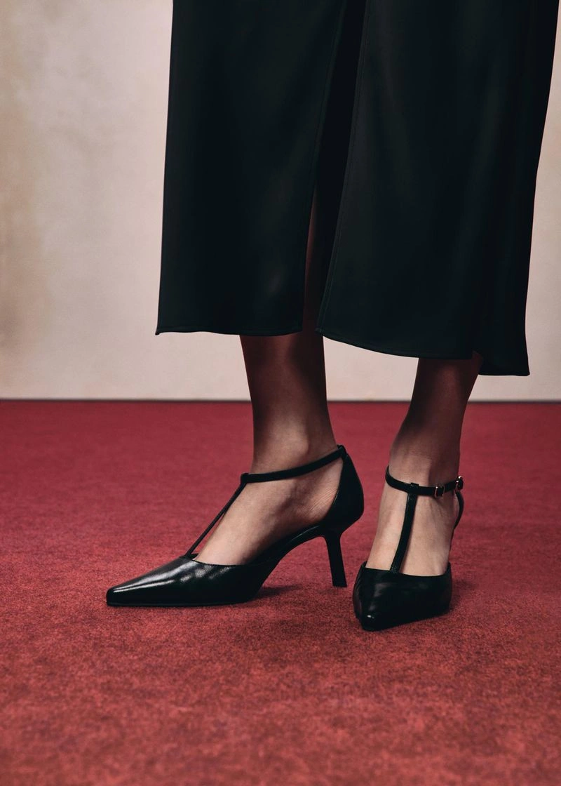 Close-up of black t-strap heels and a dark skirt standing on a red carpet against a light neutral wall.