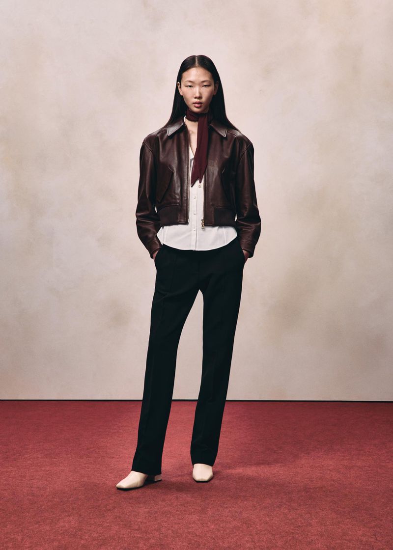 Model wearing a brown leather jacket over a white shirt and black trousers against a textured studio background