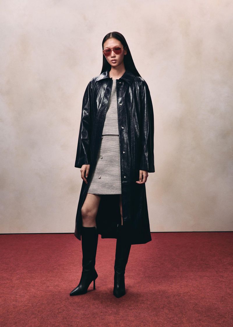 Model wearing a black glossy long coat and tall boots standing against a neutral wall over a red floor.