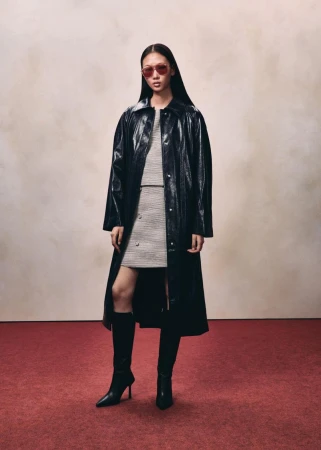 Model wearing a black glossy long coat and tall boots standing against a neutral wall over a red floor.