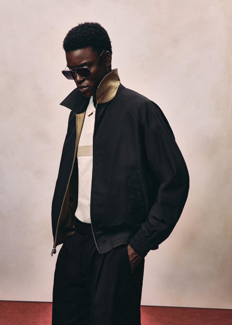 Black man wearing sunglasses, black jacket, and white polo shirt against a textured neutral background for HUGO Fall 2026.
