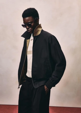Black man wearing sunglasses, black jacket, and white polo shirt against a textured neutral background for HUGO Fall 2026.