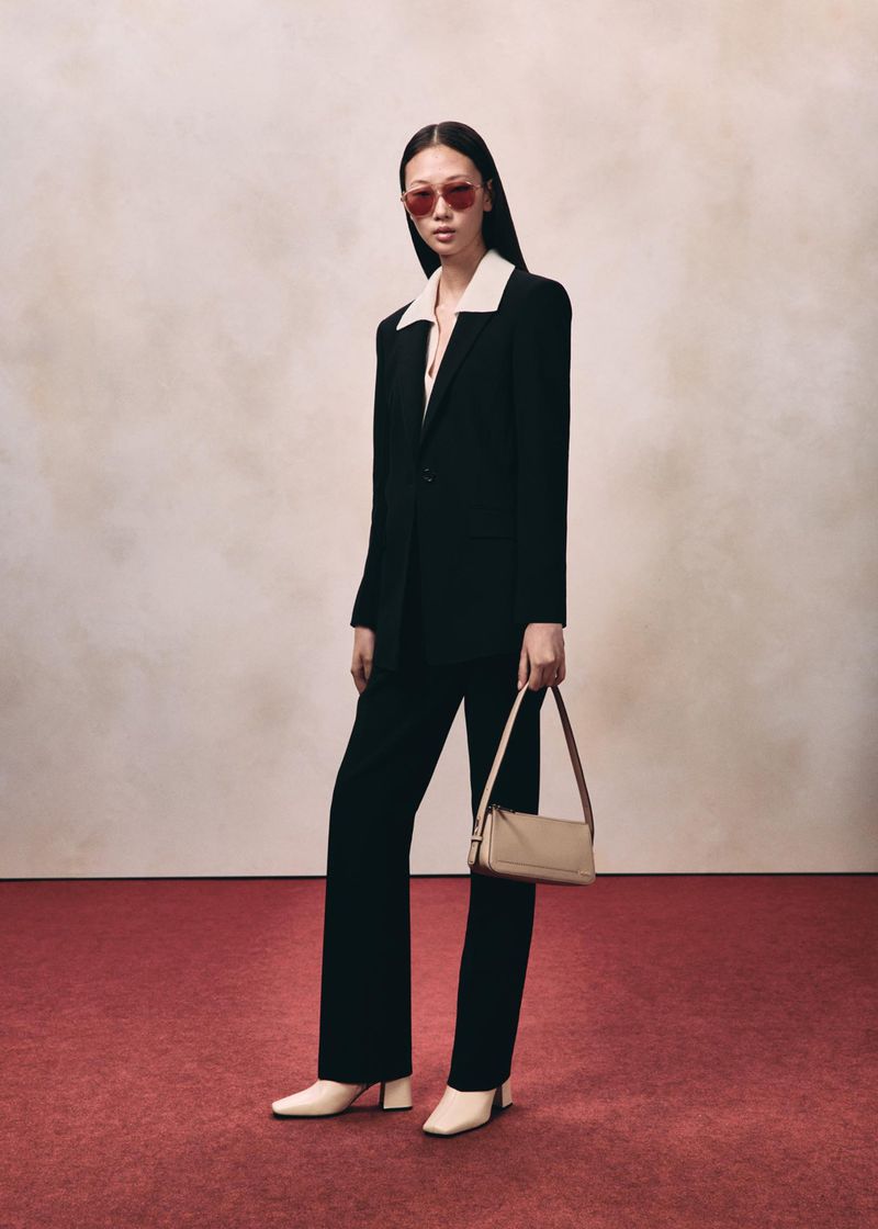 Model in black suit and red sunglasses holding a beige handbag against a textured backdrop