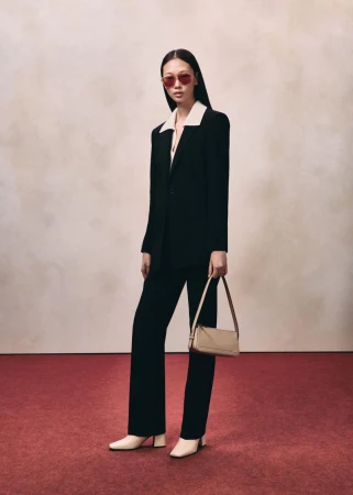 Model in black suit and red sunglasses holding a beige handbag against a textured backdrop