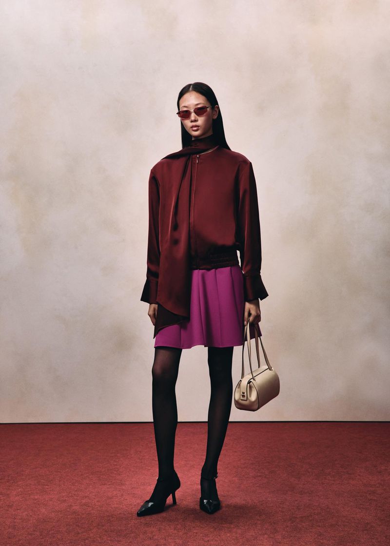 Model wearing a burgundy satin top and magenta skirt with sunglasses holding a handbag on a red carpeted floor.