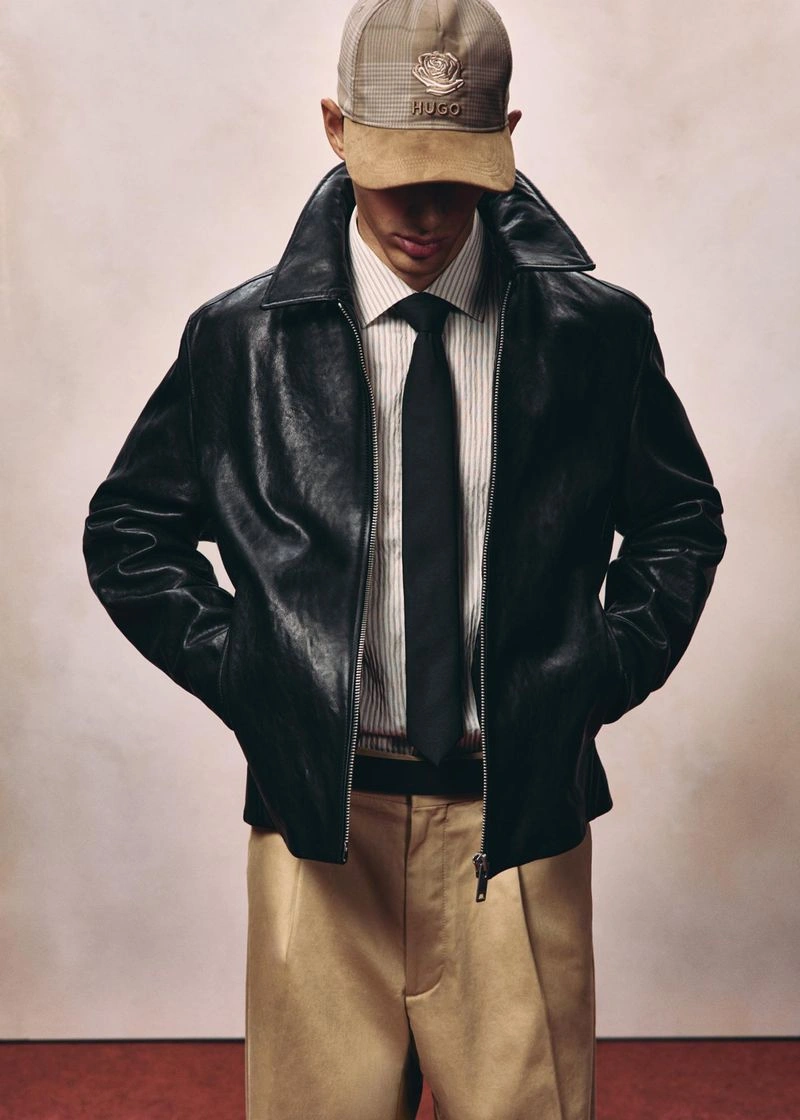 Model wearing a black leather jacket, striped shirt, tie, and plaid cap against a neutral studio backdrop