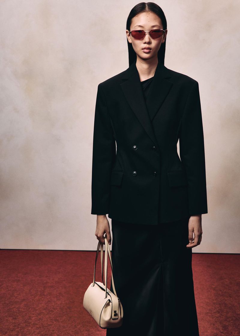 Model wearing a black blazer and sunglasses holding a light handbag against a neutral background