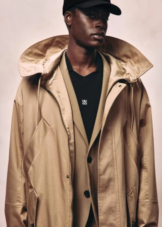 Black man wearing a black cap, layered beige coat, and jacket with black polo shirt for HUGO Red Fall 2026