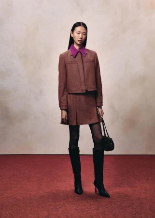 Model in brown jacket and pleated skirt holding black handbag against mottled background