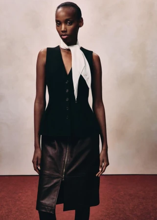 Model in black vest, white scarf, and dark skirt against neutral textured wall