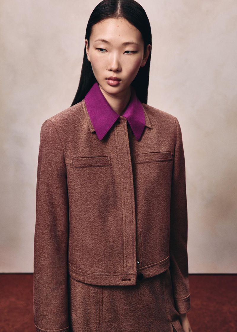 East Asian female model with long dark hair wearing a brown jacket with a purple collar against a neutral studio backdrop.