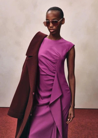 Model wearing a vibrant purple dress and tinted sunglasses, holding a burgundy coat over one shoulder against a neutral studio backdrop
