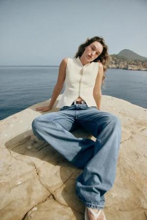 Model Jenn du Puy seated on a rocky outcrop by the sea wearing a white sleeveless top and wide-leg blue jeans for Zara.